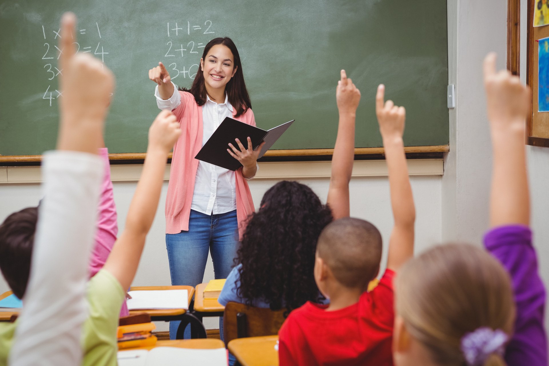 teacher standing in from of class who are raising their hands